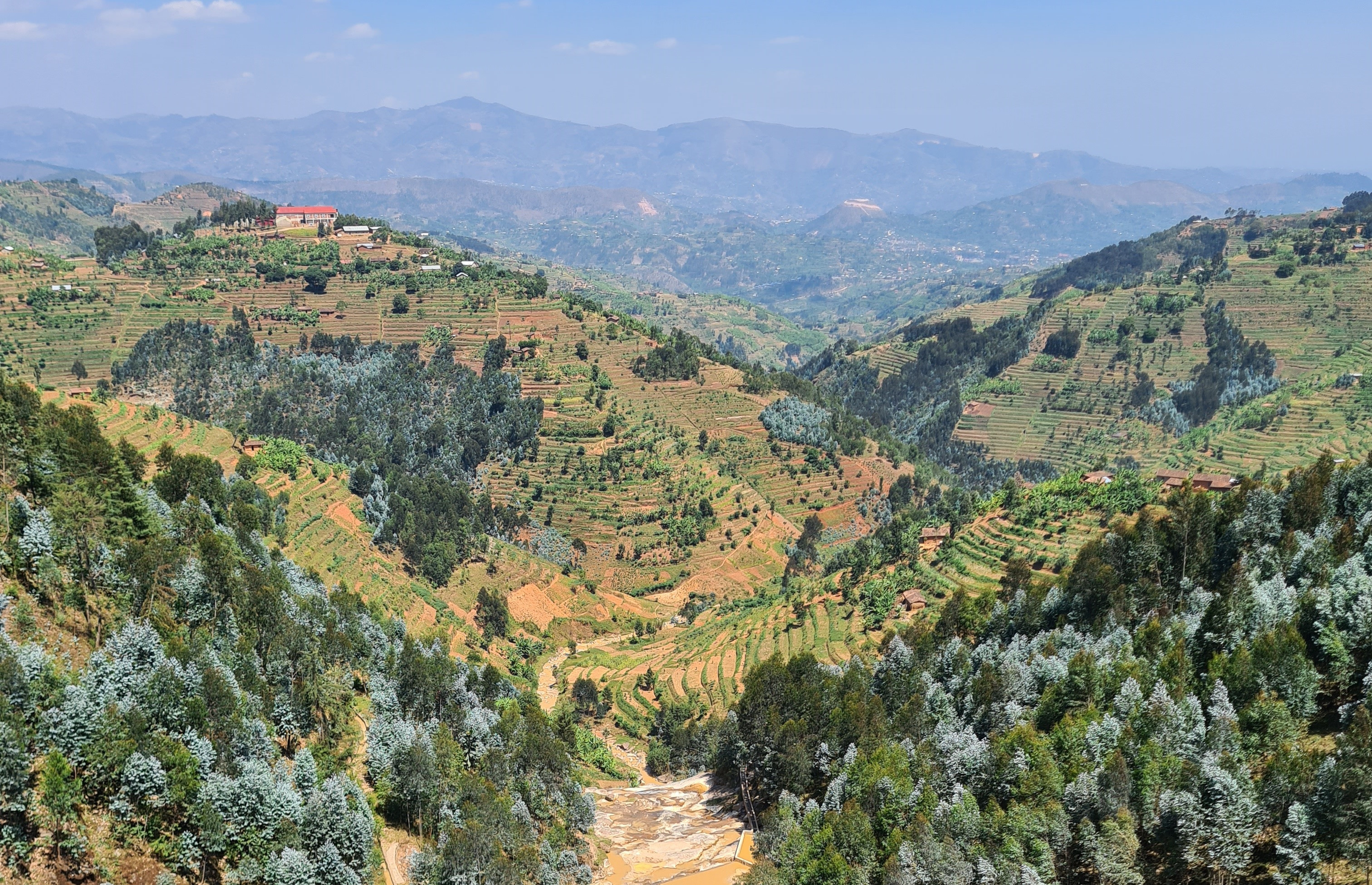Es er&ouml;ffnet sich ein weiter Blick auf eine bergige Landschaft voller B&auml;ume und landwirtschaftlicher Terrassen. In der Ferne sieht man eine Bergkette.