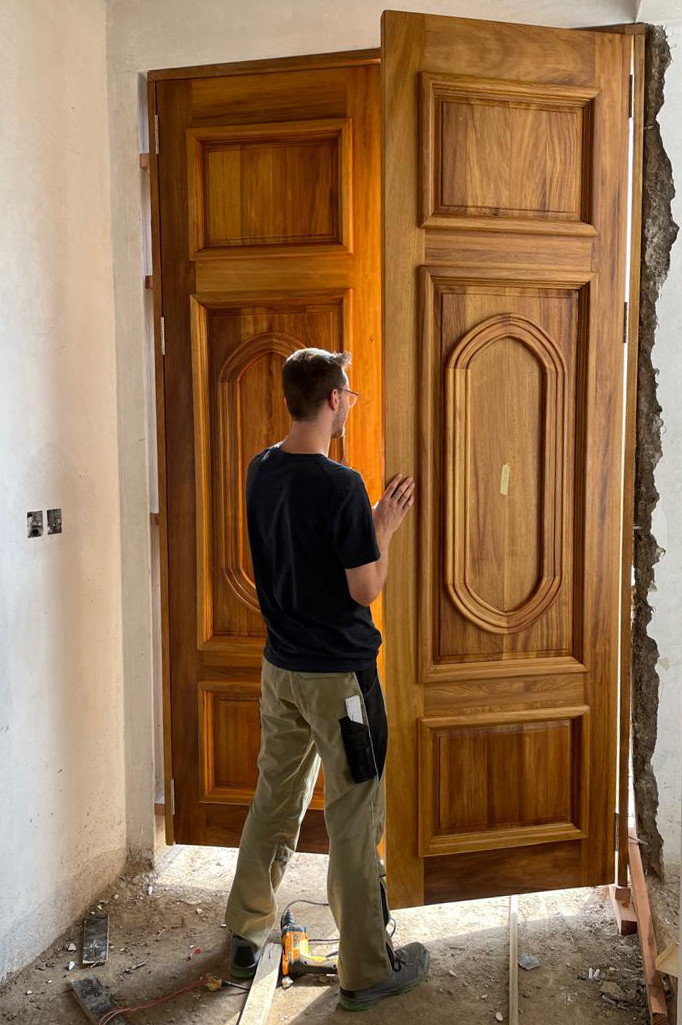 Nico arbeitet an einer gro&szlig;en Fl&uuml;gelt&uuml;r aus warmem Holz. Er steht mit dem R&uuml;cken zur Betrachterin.