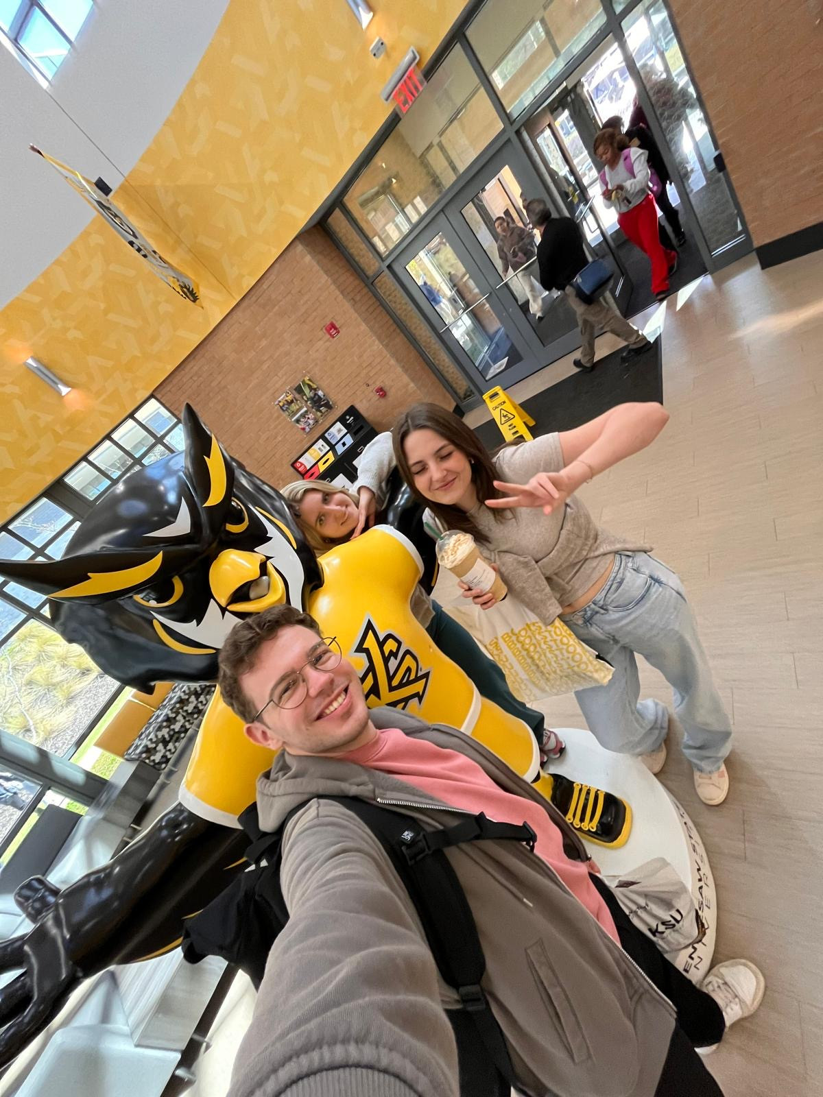 Auf dem Bild ist die Auszubildende Jessica mit anderen Alumi am College zu sehen, wie sie ein Selfie mit dem Maskottchen zu dritt machen.