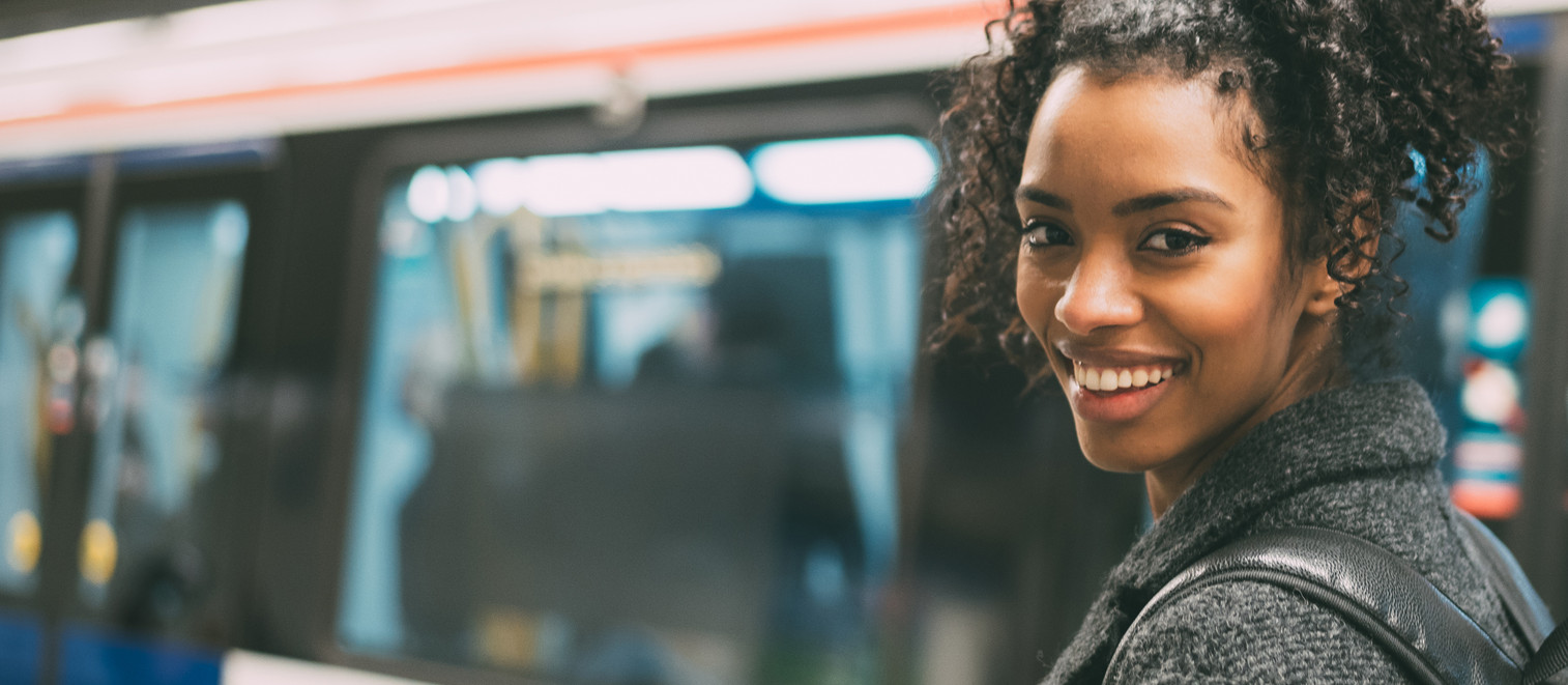 Foto: Junge Frau mit schwarzem Rucksack wendet sich lächelnd zur Kamera. Im Hintergrund sieht man verschwommen eine U-Bahn