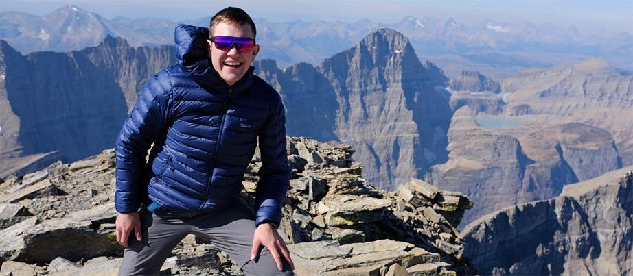 Foto: Junger Mann mit Skibrille steht auf einem Berg und lacht in die Kamera, im Hintergrund sieht man die Berge des Glacier Nationalparks