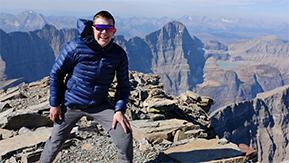 Foto: Junger Mann mit Skibrille steht auf einem Berg und lacht in die Kamera, im Hintergrund sieht man die Berge des Glacier Nationalparks