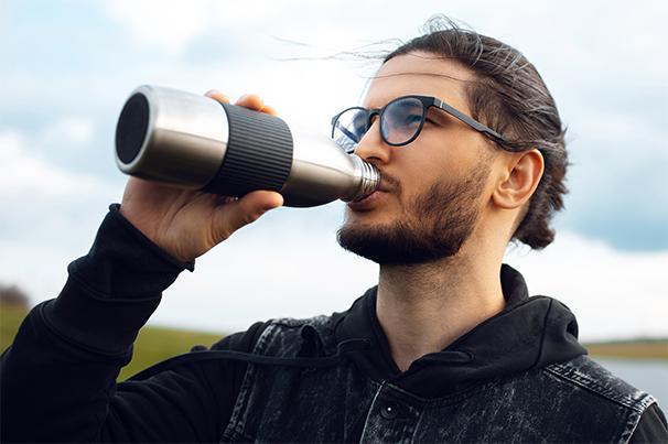 Foto: junger Mann mit Brille und Zopf trinkt aus einer Metallflasche, im Hintergrund sieht man den Himmel, grüne Hügel und Wasser
