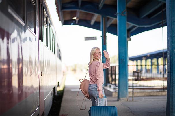 Foto: junge Frau mit Rollkoffer steht am Gleis neben dem Zug und winkt lachend in die Kamera