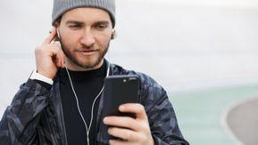 Foto: junger Mann mit grauer Mütze steckt einen Kopfhörer ins Ohr und hält sein Handy in der Hand