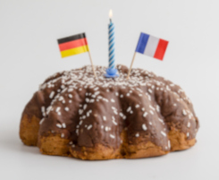 Foto eines Kuchens. Auf dem Kuchen steckt eine brennende Kerze und je eine Flagge Deutschlands und Frankreichs.