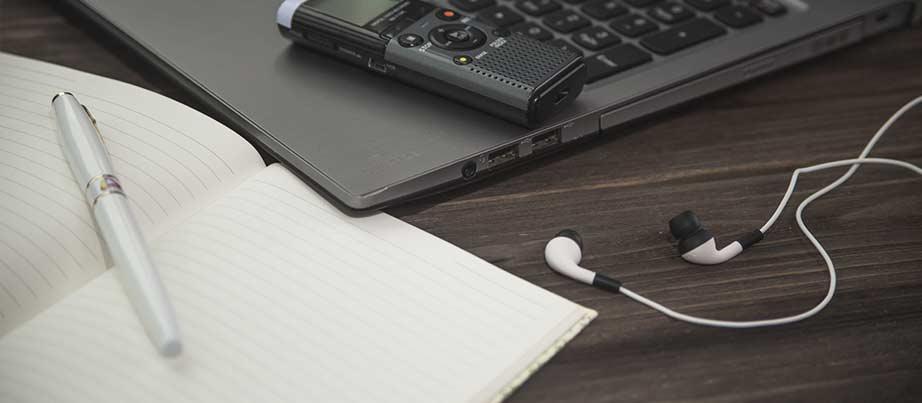 Foto von Computer, Notizbuch, Stift, Diktiergerät und Kopfhören.